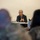 An older man with gray hair and glasses is seated at a table, appearing to address a group of people in what looks like a briefing or meeting room. Several attendees are visible in the foreground, some appearing to wear military camouflage uniforms. The speaker is holding a pen and gesturing with clasped hands, suggesting active discussion or presentation.