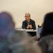 An older man with gray hair and glasses is seated at a table, appearing to address a group of people in what looks like a briefing or meeting room. Several attendees are visible in the foreground, some appearing to wear military camouflage uniforms. The speaker is holding a pen and gesturing with clasped hands, suggesting active discussion or presentation.