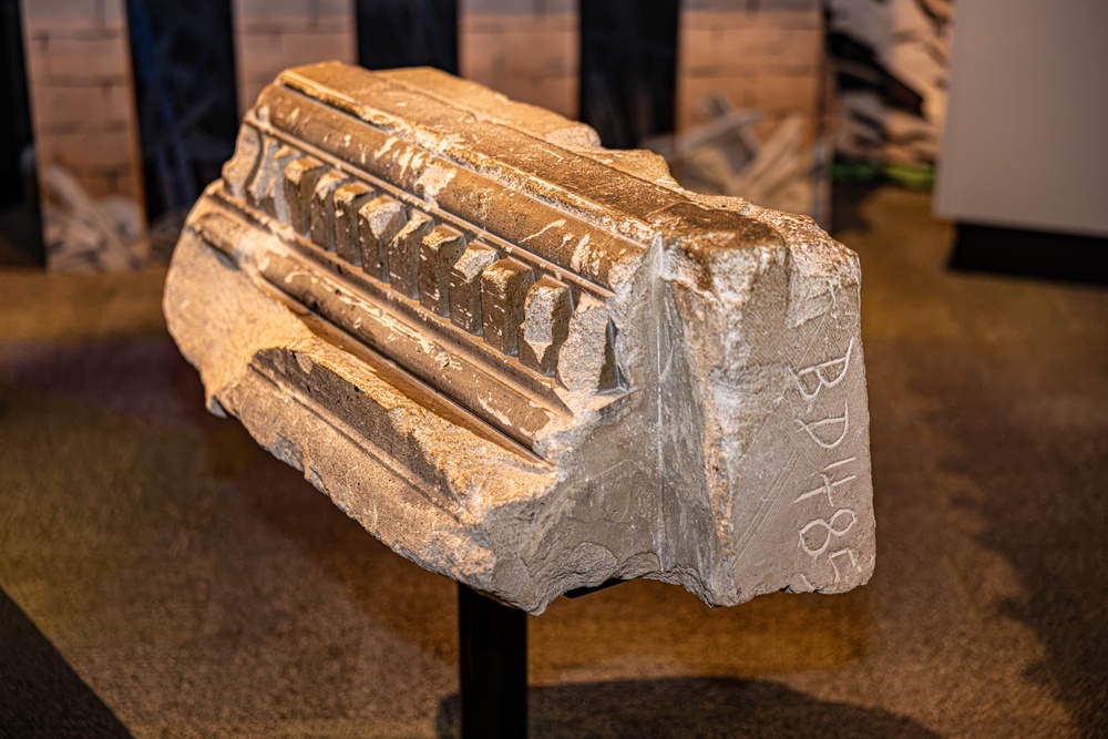A stone architectural fragment, likely from a classical or medieval building, is displayed on a metal stand in what appears to be a museum setting. The fragment features carved decorative molding with dentil and fluted details on its upper surface. An inscription or graffiti reading 'BPH+8?' is visible on the side face of the stone.