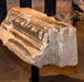 A stone architectural fragment, likely from a classical or medieval building, is displayed on a metal stand in what appears to be a museum setting. The fragment features carved decorative molding with dentil and fluted details on its upper surface. An inscription or graffiti reading 'BPH+8?' is visible on the side face of the stone.