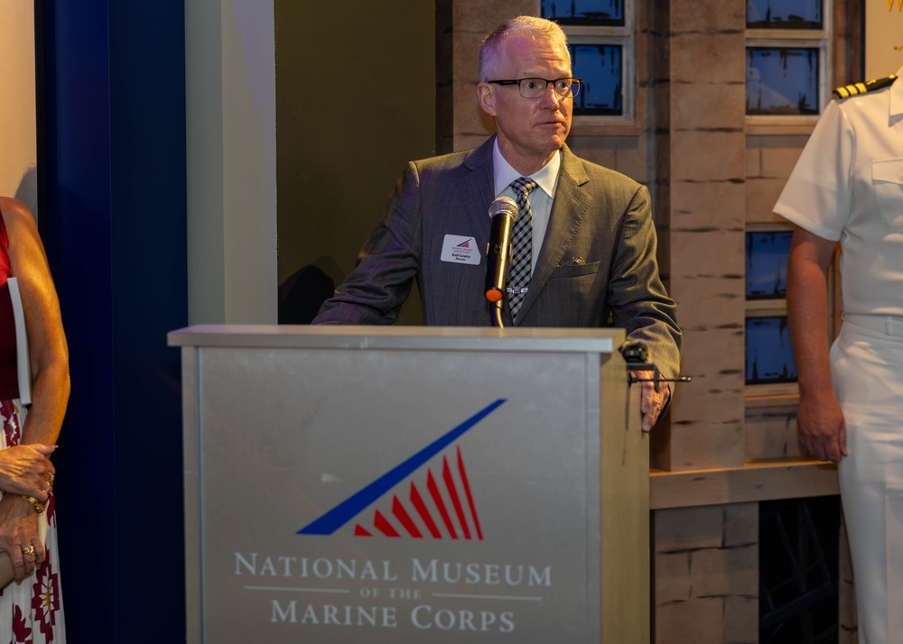 A man in a gray suit is speaking at a podium bearing the logo and name of the National Museum of the Marine Corps. He is wearing glasses, a patterned tie, and a name badge, suggesting he is an official speaker or presenter at a formal event. A person in a white military uniform is partially visible on the right side of the frame.