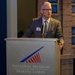 A man in a gray suit is speaking at a podium bearing the logo and name of the National Museum of the Marine Corps. He is wearing glasses, a patterned tie, and a name badge, suggesting he is an official speaker or presenter at a formal event. A person in a white military uniform is partially visible on the right side of the frame.