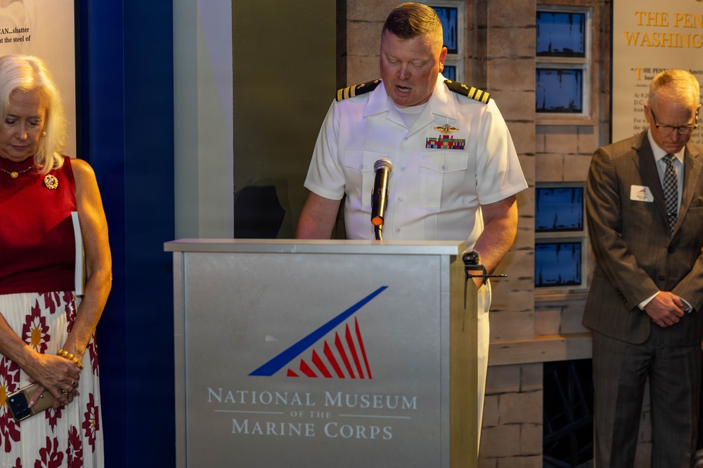 A U.S. Navy officer in white dress uniform is speaking at a podium inside the National Museum of the Marine Corps. He is flanked by a woman in a red and white outfit on the left and a man in a gray suit on the right, both with heads bowed as if in prayer or solemn reflection. The setting appears to be a formal ceremony or commemorative event at the museum.