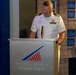 A U.S. Navy officer in white dress uniform is speaking at a podium inside the National Museum of the Marine Corps. He is flanked by a woman in a red and white outfit on the left and a man in a gray suit on the right, both with heads bowed as if in prayer or solemn reflection. The setting appears to be a formal ceremony or commemorative event at the museum.