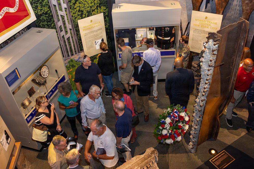 Visitors are touring a 9/11 memorial exhibition inside what appears to be a military museum, likely the National Museum of the Marine Corps or a similar institution. The exhibit features panels referencing Shanksville, Pennsylvania and the World Trade Center, New York City, along with a prominent steel beam artifact and a patriotic floral wreath. Multiple visitors of varying ages are engaged with the displays, reading panels and examining artifacts.