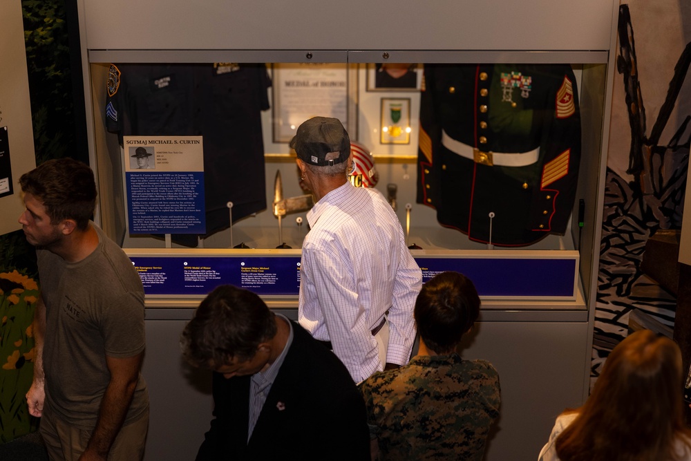 Visitors are viewing a museum display case dedicated to SGTMAJ Michael S. Curtin, featuring military and law enforcement memorabilia. The exhibit includes a Marine Corps dress uniform, an NYPD uniform, a Medal of Honor certificate, and various artifacts behind glass. Several people of different ages are gathered around the illuminated display case, reading the informational plaques.