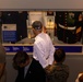Visitors are viewing a museum display case dedicated to SGTMAJ Michael S. Curtin, featuring military and law enforcement memorabilia. The exhibit includes a Marine Corps dress uniform, an NYPD uniform, a Medal of Honor certificate, and various artifacts behind glass. Several people of different ages are gathered around the illuminated display case, reading the informational plaques.