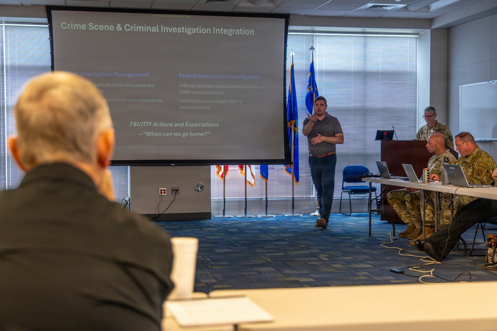 A briefing or training session is taking place in a conference room, focused on 'Crime Scene &amp; Criminal Investigation Integration' as shown on the projected presentation screen. A civilian presenter stands before the audience, which includes several individuals in military camouflage uniforms seated at a table with laptops. The setting appears to be a military or law enforcement facility, indicated by the flags and uniformed attendees.