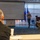 A briefing or training session is taking place in a conference room, focused on 'Crime Scene &amp; Criminal Investigation Integration' as shown on the projected presentation screen. A civilian presenter stands before the audience, which includes several individuals in military camouflage uniforms seated at a table with laptops. The setting appears to be a military or law enforcement facility, indicated by the flags and uniformed attendees.