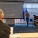 A briefing or training session is taking place in a conference room, focused on 'Crime Scene &amp; Criminal Investigation Integration' as shown on the projected presentation screen. A civilian presenter stands before the audience, which includes several individuals in military camouflage uniforms seated at a table with laptops. The setting appears to be a military or law enforcement facility, indicated by the flags and uniformed attendees.