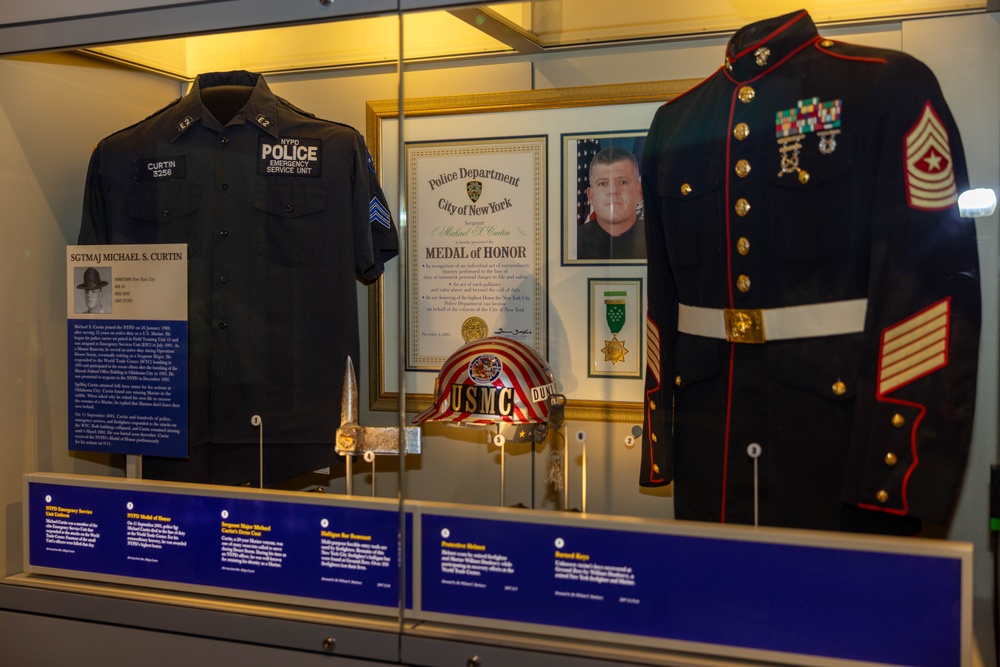 A museum display case honoring SGTMAJ Michael S. Curtin, featuring his NYPD Emergency Service Unit uniform and U.S. Marine Corps dress uniform. The exhibit includes a New York City Police Department Medal of Honor certificate, a USMC helmet, medals, ribbons, and informational plaques. The display commemorates his dual service as both a New York City police officer and a U.S. Marine.