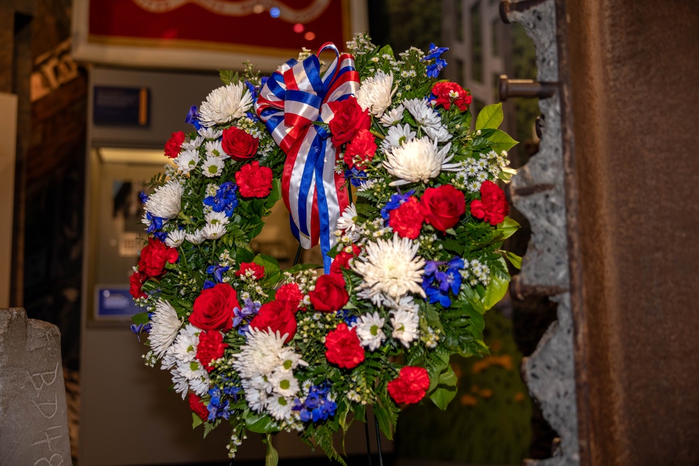A formal memorial wreath composed of red, white, and blue flowers is displayed on a stand in what appears to be an indoor museum or memorial setting. The wreath features a patriotic red, white, and blue striped ribbon bow at the top, suggesting a commemorative or ceremonial occasion. The background shows display cases and framed items consistent with a museum or memorial hall environment.