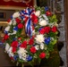 A formal memorial wreath composed of red, white, and blue flowers is displayed on a stand in what appears to be an indoor museum or memorial setting. The wreath features a patriotic red, white, and blue striped ribbon bow at the top, suggesting a commemorative or ceremonial occasion. The background shows display cases and framed items consistent with a museum or memorial hall environment.