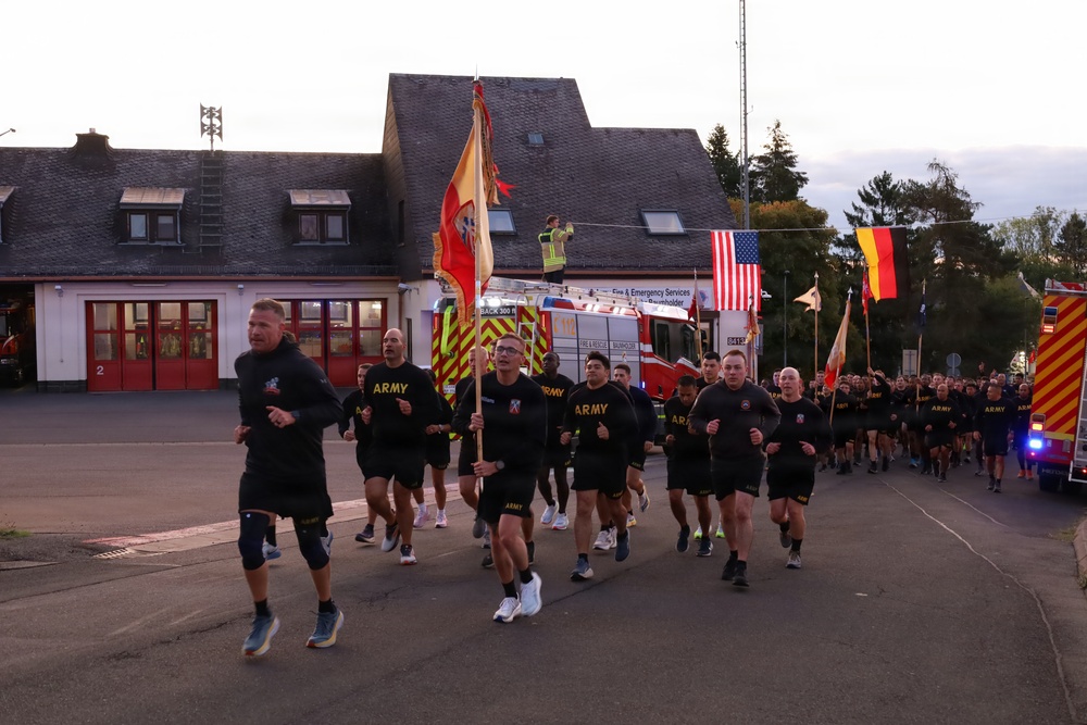 A large group of U.S. Army personnel in black PT (physical training) uniforms are conducting a formation run along a road. The run takes place near a fire and emergency services station, with an American flag and a German flag visible in the background, suggesting a U.S. military installation in Germany. Several soldiers carry military guidon flags at the front of the formation.
