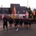 A large group of U.S. Army personnel in black PT (physical training) uniforms are conducting a formation run along a road. The run takes place near a fire and emergency services station, with an American flag and a German flag visible in the background, suggesting a U.S. military installation in Germany. Several soldiers carry military guidon flags at the front of the formation.