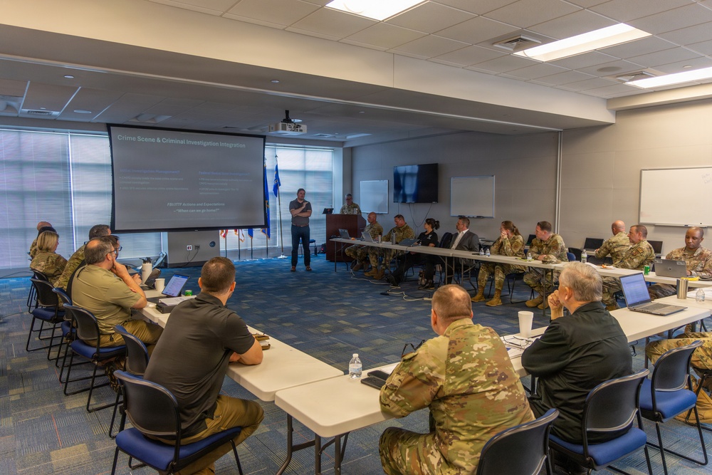 A formal military briefing or training session is taking place in a conference room, with participants seated around tables arranged in a U-shape. A presenter stands at the front near a projected screen displaying a slide titled 'Crime Scene &amp; Criminal Investigation Integration.' The majority of attendees are wearing military camouflage uniforms, with a few individuals in civilian attire.