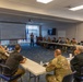A formal military briefing or training session is taking place in a conference room, with participants seated around tables arranged in a U-shape. A presenter stands at the front near a projected screen displaying a slide titled 'Crime Scene &amp; Criminal Investigation Integration.' The majority of attendees are wearing military camouflage uniforms, with a few individuals in civilian attire.