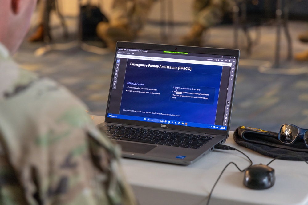 A military personnel member in camouflage uniform is viewing a training presentation on a Dell laptop. The screen displays a slide about 'Emergency Family Assistance (EFACC)' covering topics such as EFACC Activation and Communications Controls. The setting appears to be an indoor military training or briefing environment.