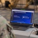 A military personnel member in camouflage uniform is viewing a training presentation on a Dell laptop. The screen displays a slide about 'Emergency Family Assistance (EFACC)' covering topics such as EFACC Activation and Communications Controls. The setting appears to be an indoor military training or briefing environment.