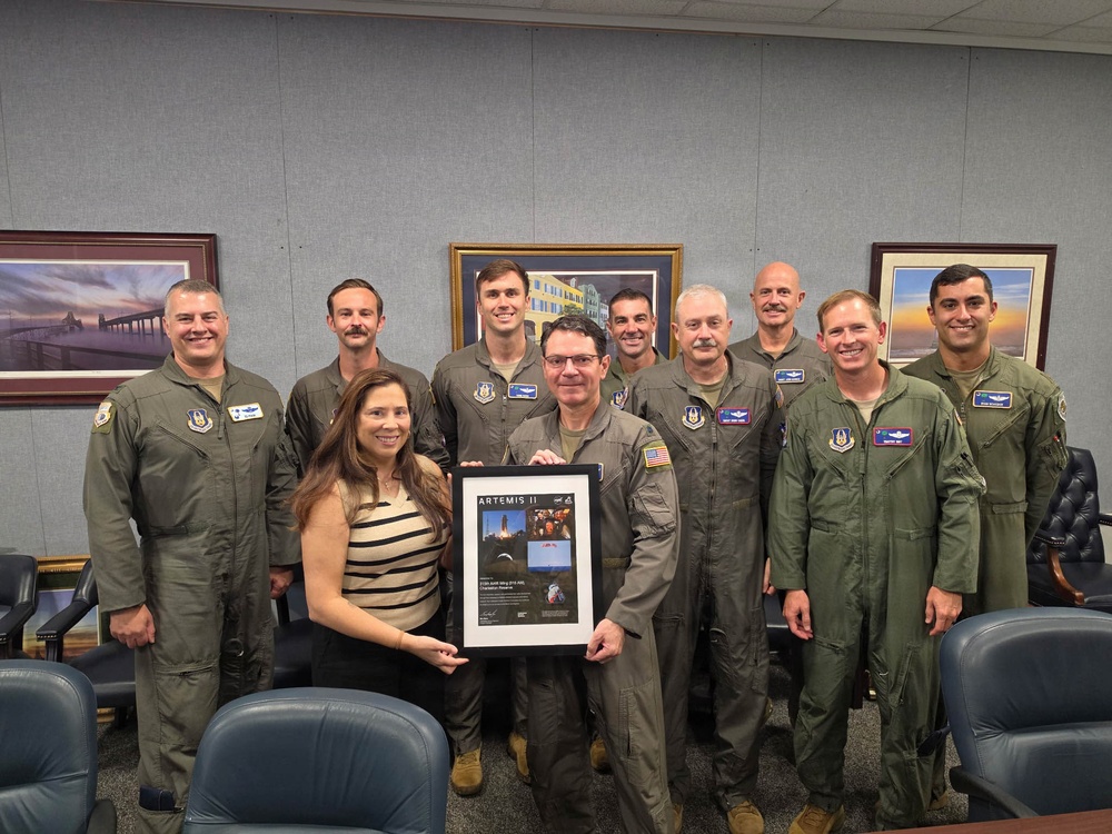 A group of approximately ten individuals, mostly wearing olive/sage green military flight suits, pose together in what appears to be a conference or briefing room. Two individuals in the center are holding a framed commemorative plaque or certificate labeled 'ARTEMIS II'. The setting suggests an official military ceremony or recognition event related to the NASA Artemis II mission.