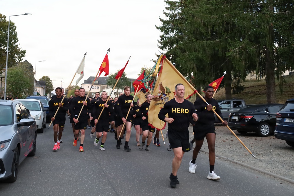 A group of U.S. Army soldiers in black physical training uniforms are running in formation down a street, carrying multiple military guidons and unit colors. The runners wear shirts labeled 'ARMY' and 'HERD', with the lead runners prominently carrying a large gold regimental flag featuring an eagle emblem. The setting appears to be a European town, likely a military installation or surrounding community, with parked cars lining both sides of the road.