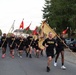 A group of U.S. Army soldiers in black physical training uniforms are running in formation down a street, carrying multiple military guidons and unit colors. The runners wear shirts labeled 'ARMY' and 'HERD', with the lead runners prominently carrying a large gold regimental flag featuring an eagle emblem. The setting appears to be a European town, likely a military installation or surrounding community, with parked cars lining both sides of the road.