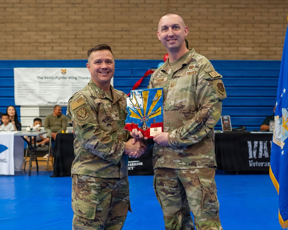Two U.S. Air Force personnel in OCP (Operational Camouflage Pattern) uniforms are shaking hands while jointly holding a colorful award or plaque during what appears to be a military recognition ceremony. The event is taking place in a gymnasium or indoor sports facility, with a banner referencing the '944th Fighter Wing' visible in the background. The award features blue, yellow, and red colors consistent with Arizona state imagery.