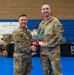Two U.S. Air Force personnel in OCP (Operational Camouflage Pattern) uniforms are shaking hands while jointly holding a colorful award or plaque during what appears to be a military recognition ceremony. The event is taking place in a gymnasium or indoor sports facility, with a banner referencing the '944th Fighter Wing' visible in the background. The award features blue, yellow, and red colors consistent with Arizona state imagery.