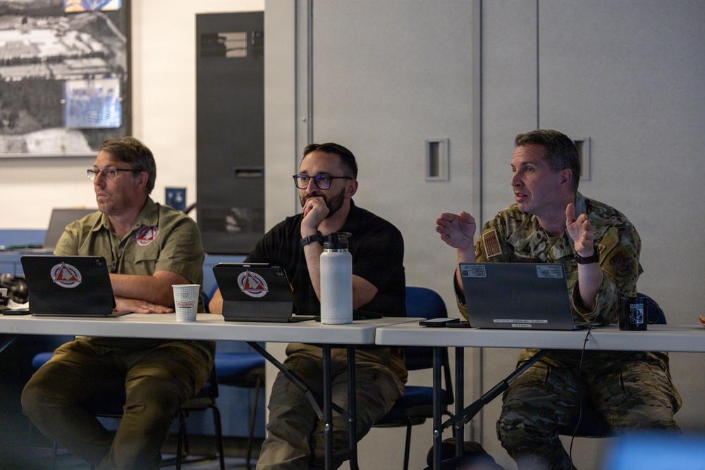 Three individuals are seated at a folding table in what appears to be a briefing or meeting room. The person on the right is wearing military camouflage (OCP/MultiCam) uniform and is actively speaking with hand gestures, while the other two listen attentively. Multiple tablets and a laptop are visible on the table, all bearing matching red insignia stickers.