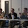 Three individuals are seated at a folding table in what appears to be a briefing or meeting room. The person on the right is wearing military camouflage (OCP/MultiCam) uniform and is actively speaking with hand gestures, while the other two listen attentively. Multiple tablets and a laptop are visible on the table, all bearing matching red insignia stickers.
