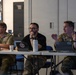 Three individuals are seated at a folding table in what appears to be a briefing or meeting room. The person on the right is wearing military camouflage (OCP/MultiCam) uniform and is actively speaking with hand gestures, while the other two listen attentively. Multiple tablets and a laptop are visible on the table, all bearing matching red insignia stickers.