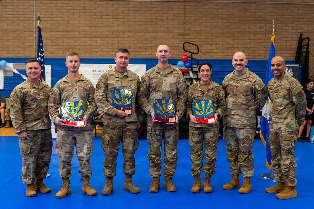Seven U.S. Air Force personnel in OCP (Operational Camouflage Pattern) uniforms pose for a group photo at what appears to be an awards ceremony. Four of the individuals are holding decorative plaques featuring what appears to be the Arizona state flag design in blue, gold, and red. The event appears to be associated with the 944th Fighter Wing based on a visible banner in the background.