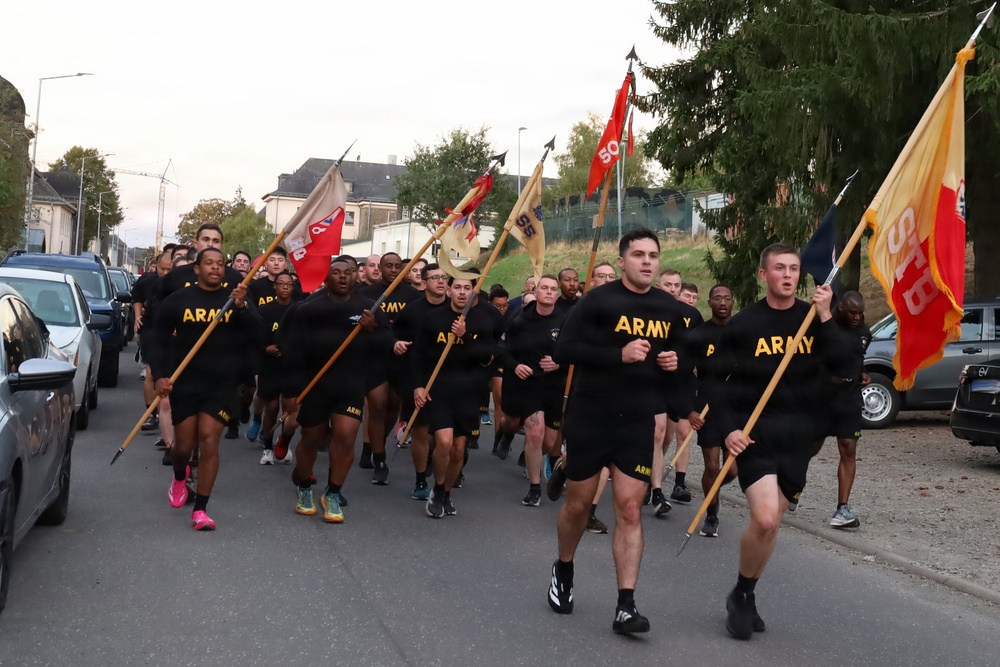 A large group of U.S. Army soldiers in black physical training (PT) uniforms are running in formation down a street, carrying unit guidon flags. The run appears to be a formation PT run or commemorative run, taking place in what appears to be a European town based on the architecture. Multiple unit guidons are visible, suggesting participation from several different Army units.