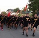 A large group of U.S. Army soldiers in black physical training (PT) uniforms are running in formation down a street, carrying unit guidon flags. The run appears to be a formation PT run or commemorative run, taking place in what appears to be a European town based on the architecture. Multiple unit guidons are visible, suggesting participation from several different Army units.