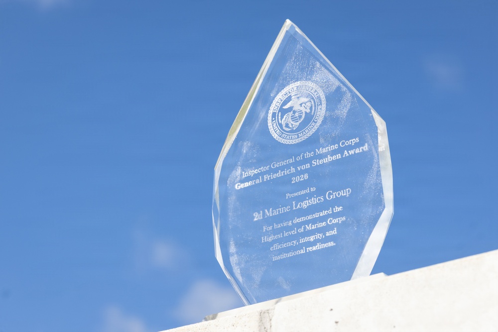 A clear glass award trophy is displayed against a bright blue sky with light clouds. The award is the 'General Friedrich von Steuben Award' for 2026, presented by the Inspector General of the Marine Corps to the 2d Marine Logistics Group. The engraved text recognizes the unit for demonstrating the highest level of Marine Corps efficiency, integrity, and institutional readiness.