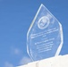 A clear glass award trophy is displayed against a bright blue sky with light clouds. The award is the 'General Friedrich von Steuben Award' for 2026, presented by the Inspector General of the Marine Corps to the 2d Marine Logistics Group. The engraved text recognizes the unit for demonstrating the highest level of Marine Corps efficiency, integrity, and institutional readiness.