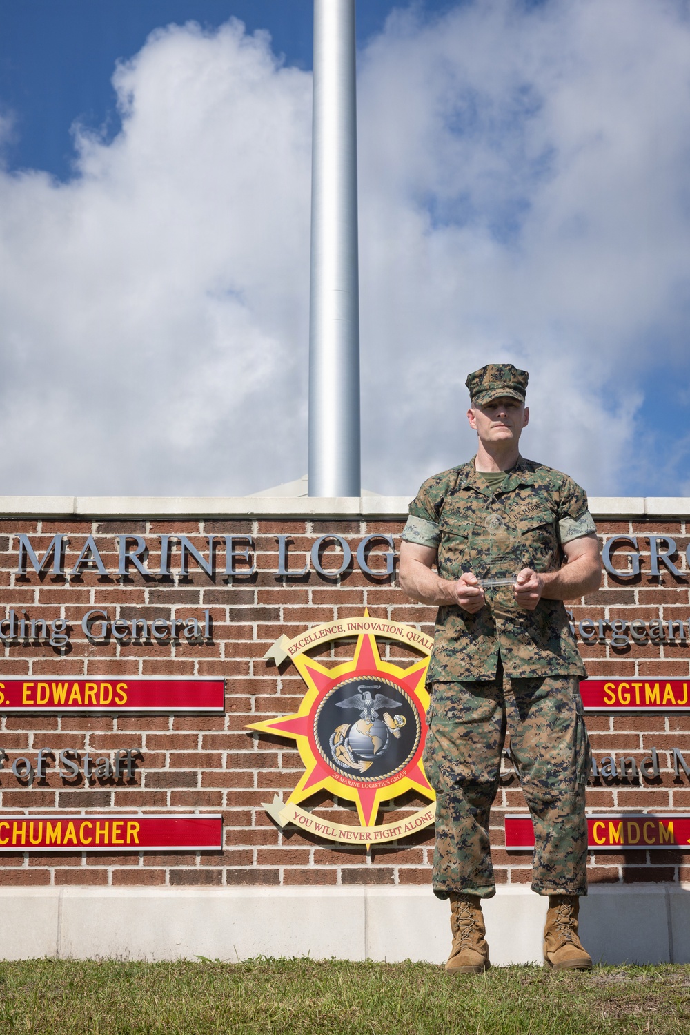 A U.S. Marine in woodland MARPAT camouflage uniform stands at ease in front of a brick command sign for the Marine Logistics Group. The sign displays unit leadership names including 'S. Edwards' and 'Schumacher,' along with the Marine Logistics Group emblem bearing the motto 'You Will Never Fight Alone.' A flagpole rises behind the subject against a partly cloudy sky.