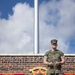 A U.S. Marine in woodland MARPAT camouflage uniform stands at ease in front of a brick command sign for the Marine Logistics Group. The sign displays unit leadership names including 'S. Edwards' and 'Schumacher,' along with the Marine Logistics Group emblem bearing the motto 'You Will Never Fight Alone.' A flagpole rises behind the subject against a partly cloudy sky.
