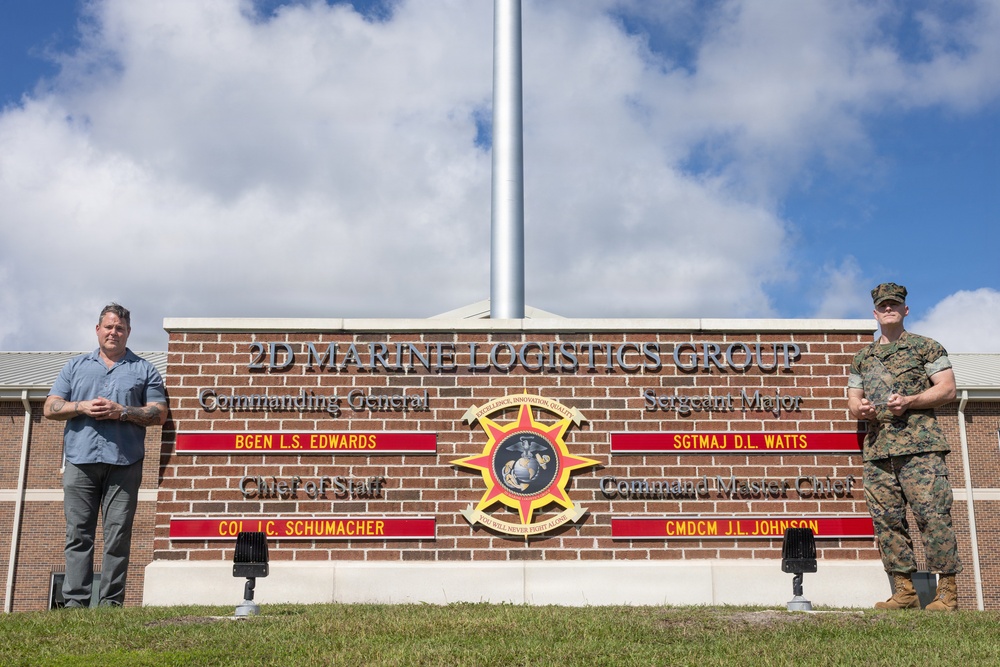 Two individuals stand in front of the 2D Marine Logistics Group headquarters sign at what appears to be a U.S. Marine Corps installation. One person is in civilian attire (blue shirt, gray pants) on the left, while the other is in Marine Corps MARPAT camouflage uniform on the right. The brick sign displays command leadership names including Commanding General BGEN L.S. Edwards, Sergeant Major SGTMAJ D.L. Watts, Chief of Staff COL J.C. Schumacher, and Command Master Chief CMDCM J.L. Johnson.
