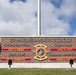 Two individuals stand in front of the 2D Marine Logistics Group headquarters sign at what appears to be a U.S. Marine Corps installation. One person is in civilian attire (blue shirt, gray pants) on the left, while the other is in Marine Corps MARPAT camouflage uniform on the right. The brick sign displays command leadership names including Commanding General BGEN L.S. Edwards, Sergeant Major SGTMAJ D.L. Watts, Chief of Staff COL J.C. Schumacher, and Command Master Chief CMDCM J.L. Johnson.