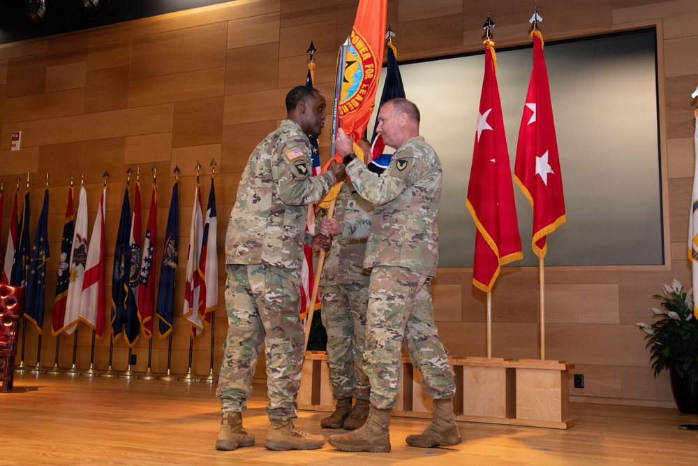 A U.S. Army change of command ceremony is taking place on a stage, with two soldiers exchanging a unit guidon flag. Red general officer flags with stars are visible in the background, indicating senior military leadership is present. Multiple state and organizational flags line the back wall of what appears to be a formal auditorium or ceremonial hall.
