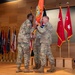 A U.S. Army change of command ceremony is taking place on a stage, with two soldiers exchanging a unit guidon flag. Red general officer flags with stars are visible in the background, indicating senior military leadership is present. Multiple state and organizational flags line the back wall of what appears to be a formal auditorium or ceremonial hall.
