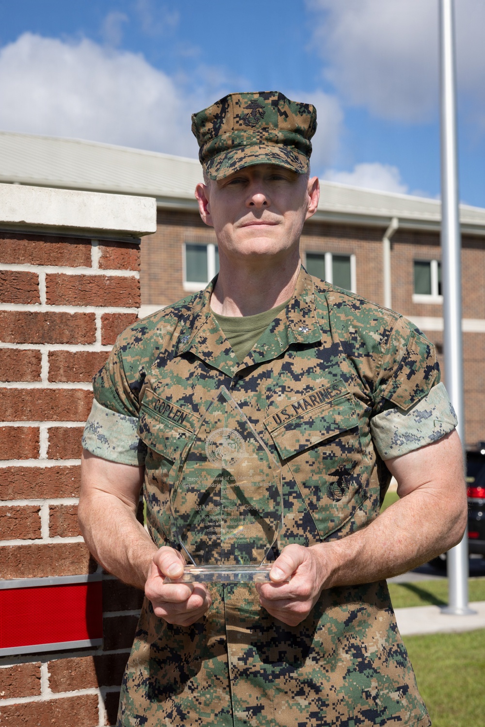 A U.S. Marine in MARPAT woodland camouflage uniform is holding a clear glass/acrylic award plaque while standing outdoors in front of a brick building. The Marine's name tape reads 'COPLEN' and the branch tape reads 'U.S. MARINES'. The award plaque appears to reference the Inspector General of the Marine Corps and the 2d Marine Logistics Group.