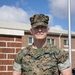 A U.S. Marine in MARPAT woodland camouflage uniform is holding a clear glass/acrylic award plaque while standing outdoors in front of a brick building. The Marine's name tape reads 'COPLEN' and the branch tape reads 'U.S. MARINES'. The award plaque appears to reference the Inspector General of the Marine Corps and the 2d Marine Logistics Group.