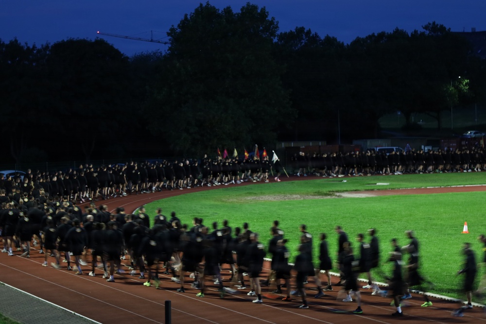 A large group of people dressed uniformly in black clothing are marching or running along a red athletic track at dusk or early morning. The scene appears to be a military or organized group physical training event, with hundreds of participants moving in formation around the track. Several colorful flags or guidons are visible among the formation, suggesting a military unit run or ceremonial march.