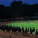 A large group of people dressed uniformly in black clothing are marching or running along a red athletic track at dusk or early morning. The scene appears to be a military or organized group physical training event, with hundreds of participants moving in formation around the track. Several colorful flags or guidons are visible among the formation, suggesting a military unit run or ceremonial march.