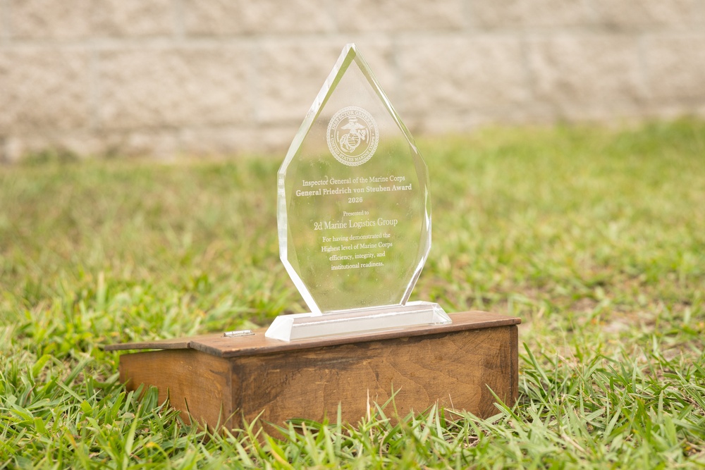 A clear glass/crystal award trophy in a diamond shape sits on a wooden base placed on grass outdoors. The award is from the Inspector General of the Marine Corps, specifically the 'General Friedrich von Steuben Award 2026,' presented to the 2d Marine Logistics Group. The engraving recognizes the unit for demonstrating the highest level of Marine Corps efficiency, integrity, and institutional readiness.