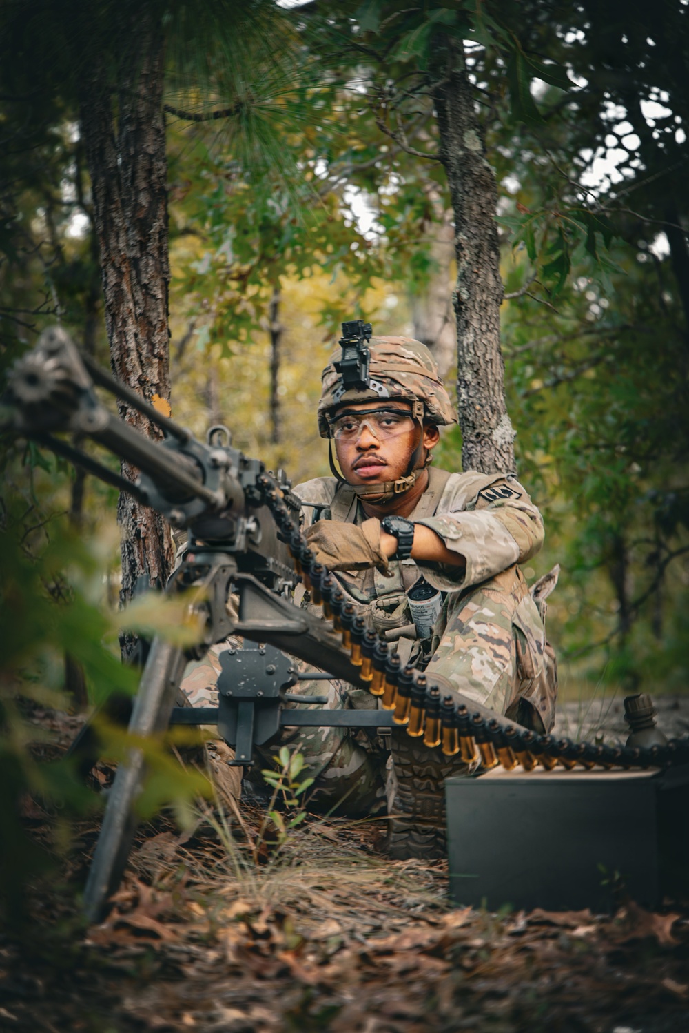 A U.S. Army soldier in full OCP (Operational Camouflage Pattern) uniform is positioned in a wooded area behind a crew-served machine gun mounted on a tripod. The soldier is handling an ammunition belt fed into the weapon while maintaining situational awareness, suggesting a field training exercise. The setting appears to be a forested training environment with mixed deciduous and coniferous trees.