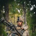 A U.S. Army soldier in full OCP (Operational Camouflage Pattern) uniform is positioned in a wooded area behind a crew-served machine gun mounted on a tripod. The soldier is handling an ammunition belt fed into the weapon while maintaining situational awareness, suggesting a field training exercise. The setting appears to be a forested training environment with mixed deciduous and coniferous trees.
