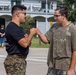 Two young men are engaged in a friendly handshake or greeting gesture in an outdoor setting. One individual is wearing military camouflage pants and a black t-shirt, while the other wears a graphic t-shirt and camouflage-patterned pants. They appear to be interacting positively in what looks like a public or institutional outdoor area.